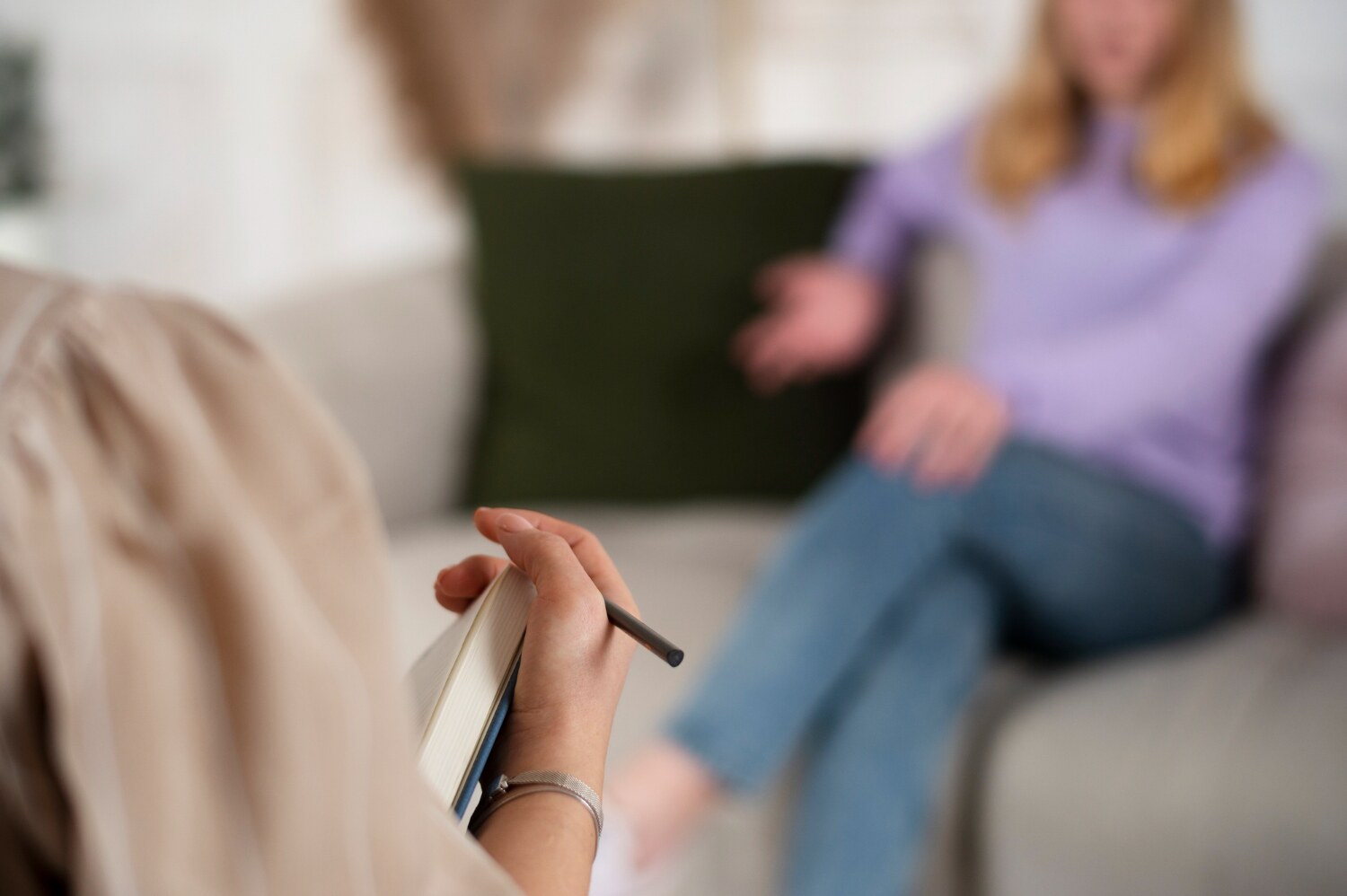 A person holds a notebook and pen, sitting opposite a blurred figure on a couch.